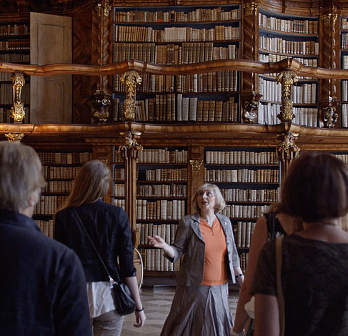 Eine Stadtführerin präsentiert Gästen die historische Provinzialbibliothek in Neuburg an der Donau. Im Hintergrund sind die prunkvollen rokoko Bücherregale zu sehen.