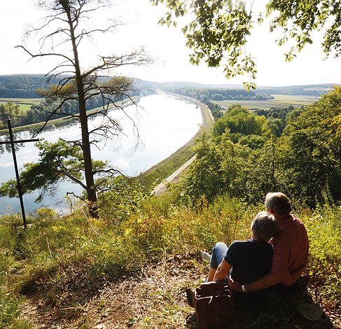 Ein Ehepaar sitzt im grünen Gras und genießt den Blick auf die sonnige Donau.